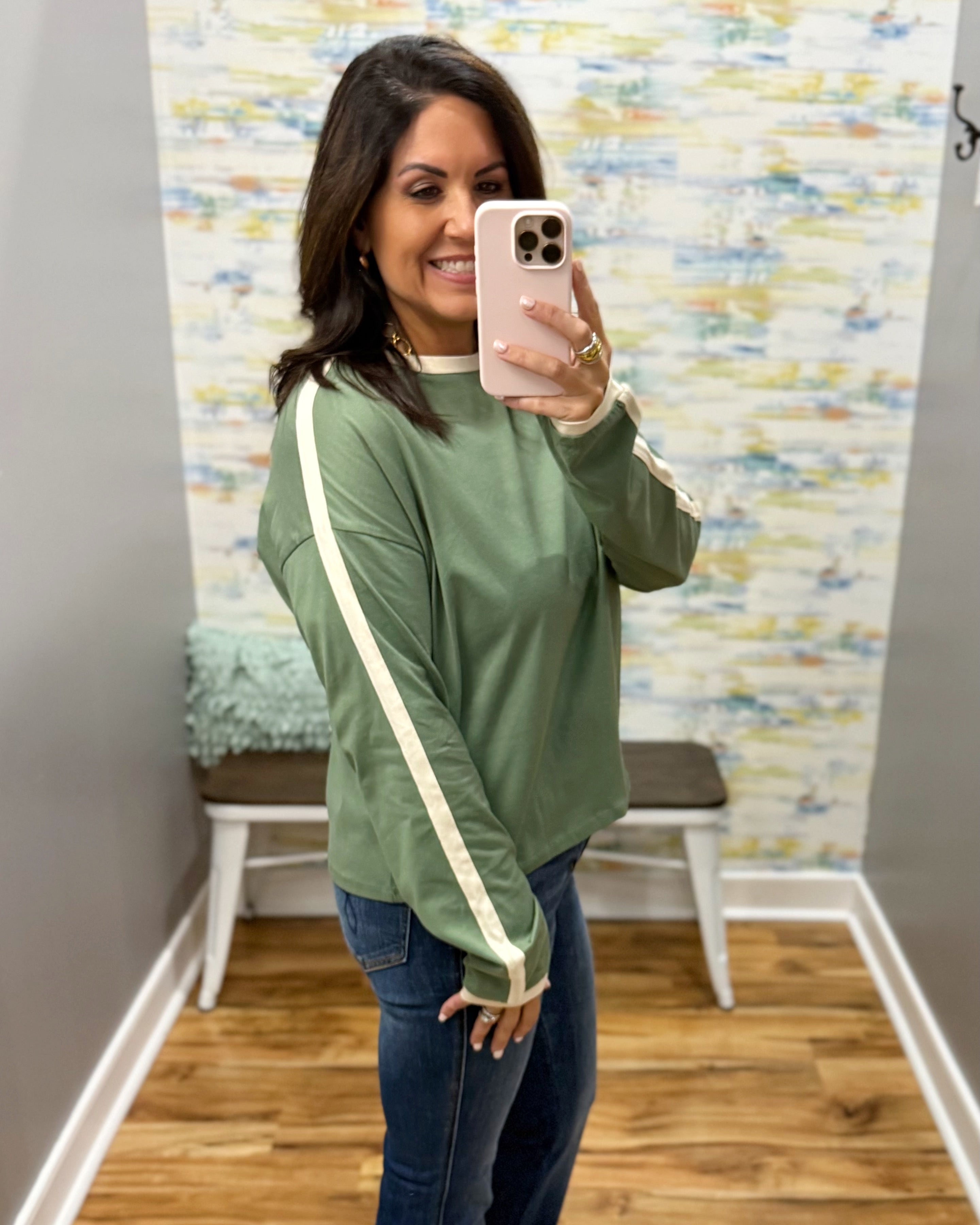 A woman takes a mirror selfie wearing an olive green long-sleeve sweatshirt with cream-colored stripes down the sleeves, paired with blue jeans.