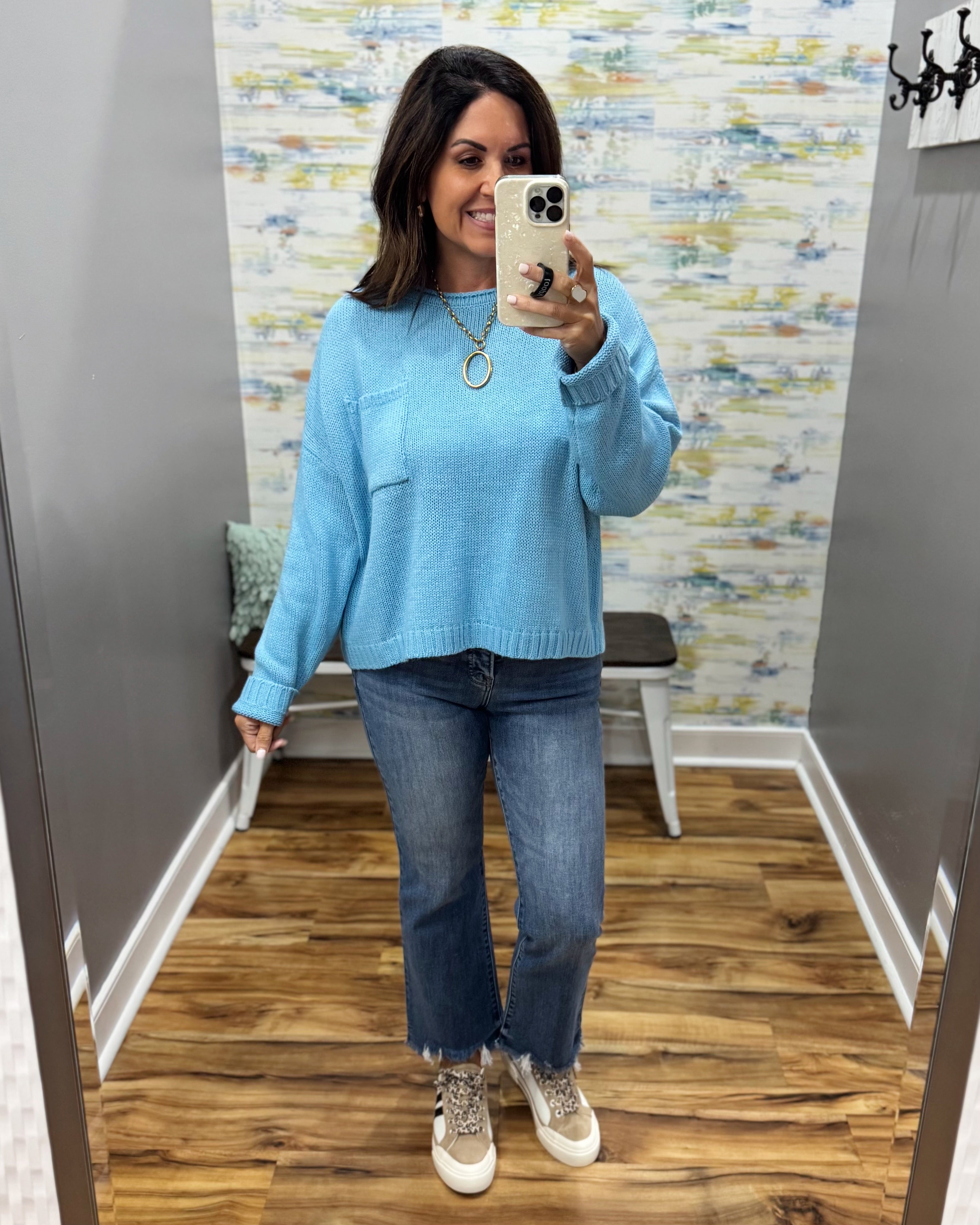 A woman takes a mirror selfie in a fitting room wearing a light blue knit crewneck sweater, blue jeans, and beige sneakers.