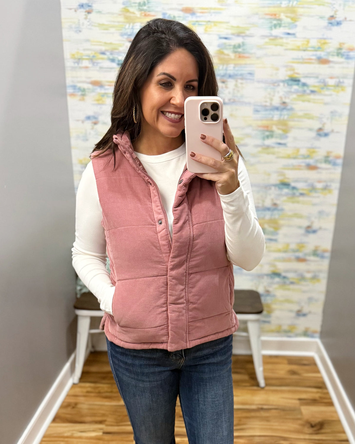 A woman takes a mirror selfie wearing a dusty pink sleeveless quilted vest over a white long-sleeve top and blue jeans. The background shows a gray wall, a patterned wallpaper, and a wooden floor.