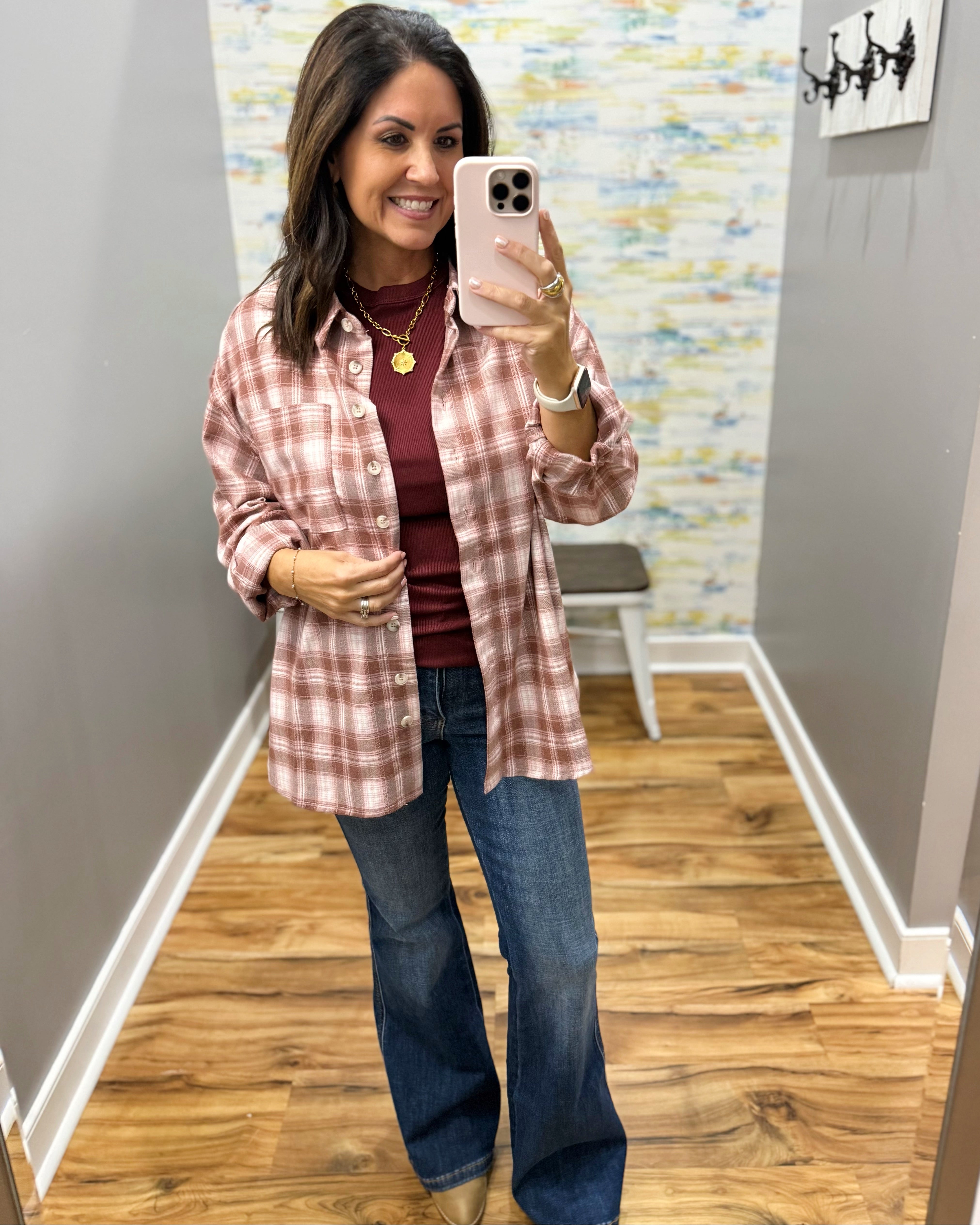 A woman takes a mirror selfie wearing a dusty pink and white plaid button-down shirt over a burgundy top, paired with blue flare jeans.