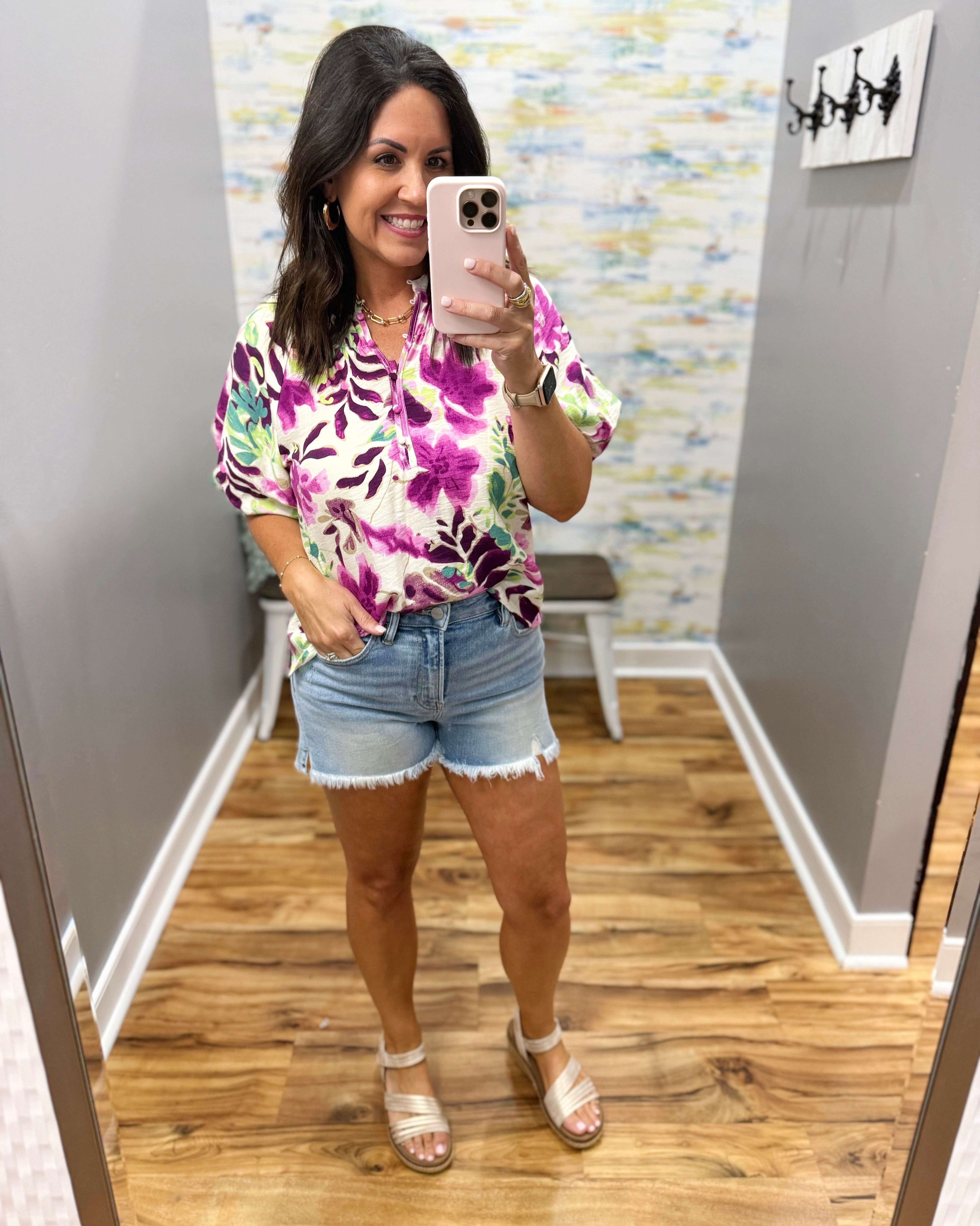 Woman taking a mirror selfie wearing a colorful floral shirt and denim shorts in a room with a patterned wall.