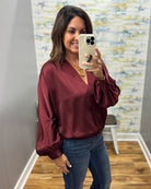 A woman takes a mirror selfie wearing a burgundy satin blouse with voluminous long sleeves and blue jeans in a fitting room.