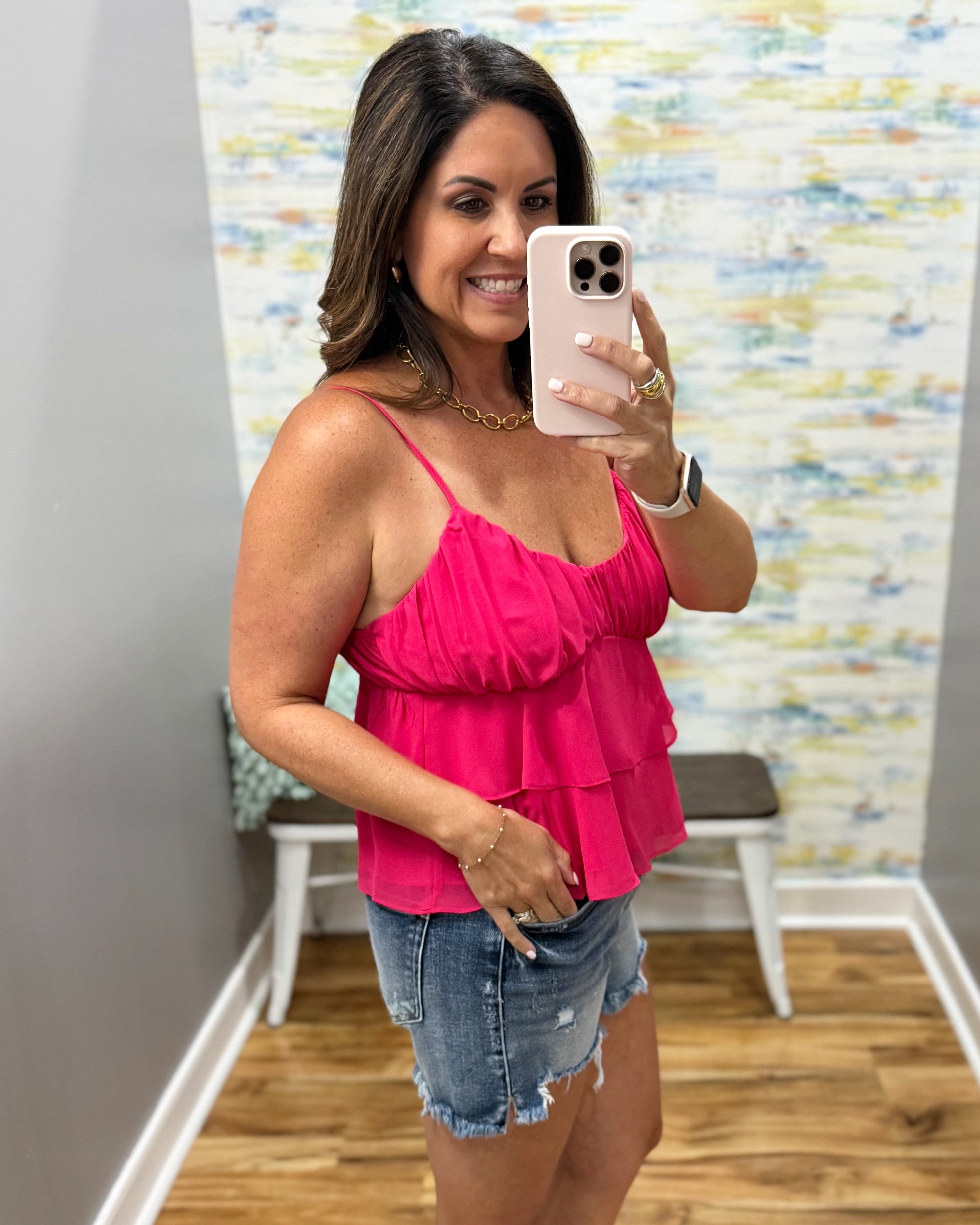 Woman taking a mirror selfie wearing a pink top and denim shorts in a room with a colorful wall.