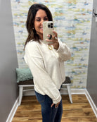 A woman takes a mirror selfie wearing a cream-colored knit zip-up sweater and dark blue jeans in a room with a patterned wallpaper background.