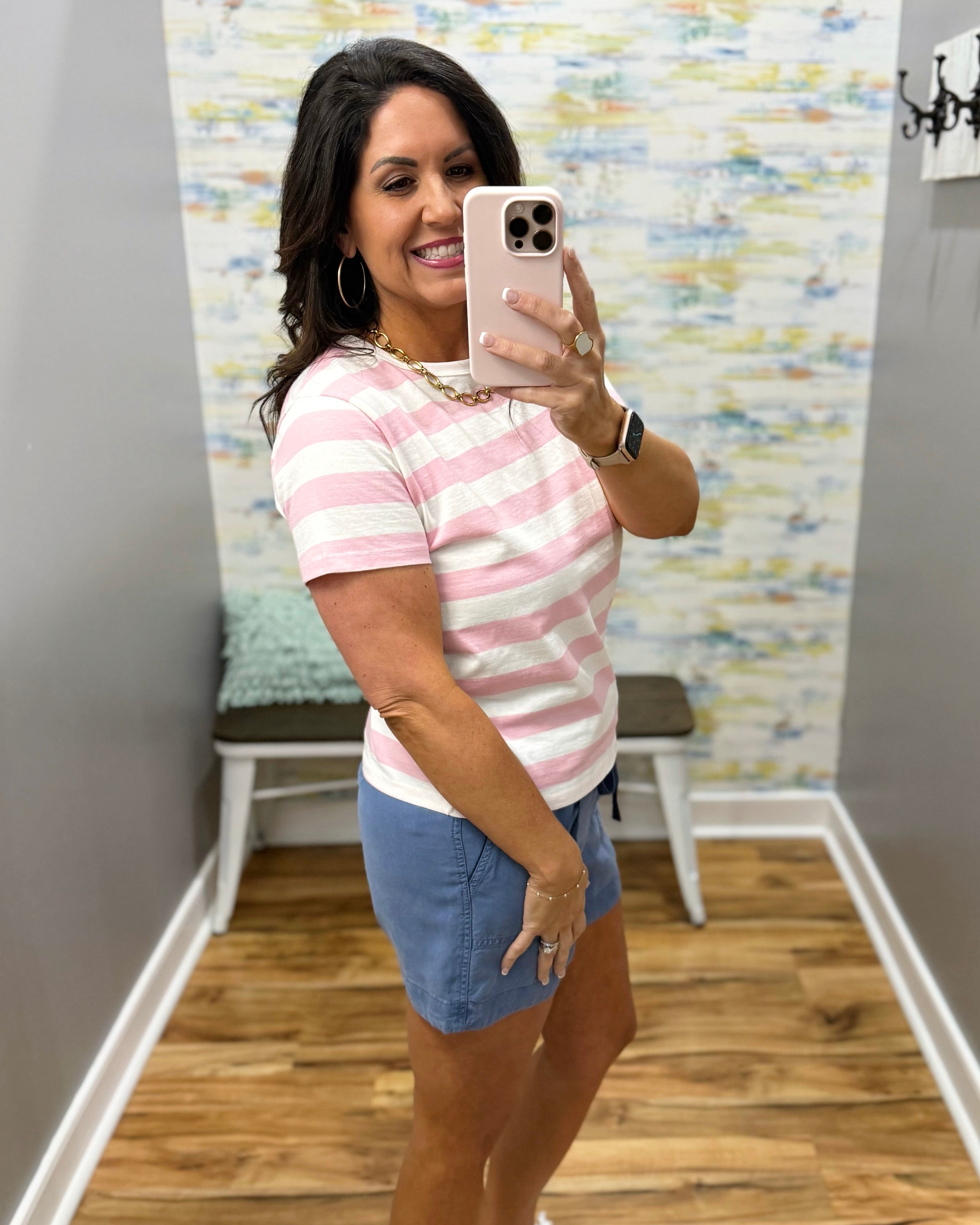 Woman taking a mirror selfie wearing a pink and white striped shirt and blue shorts.