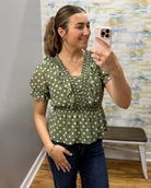 A woman takes a mirror selfie wearing an olive green short-sleeve blouse with white polka dots and ruffle details, paired with dark blue jeans.