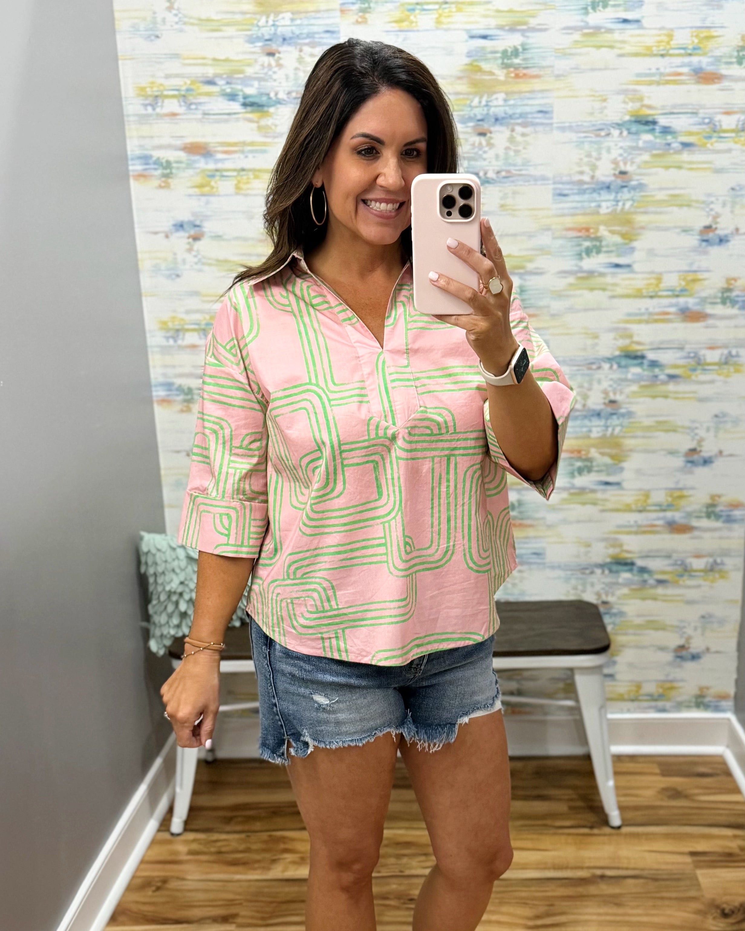 Woman taking a mirror selfie wearing a pink and green patterned shirt and denim shorts in a room with colorful wall art.