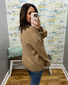 A woman takes a mirror selfie wearing a loose-fit, light brown knit sweater and blue jeans in a home setting with a patterned wallpaper background.