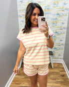 A woman takes a mirror selfie wearing a matching two-piece pajama set with light pink and white horizontal stripes. The set includes a short-sleeve crewneck t-shirt and matching shorts, shown in a home or boutique fitting room environment.