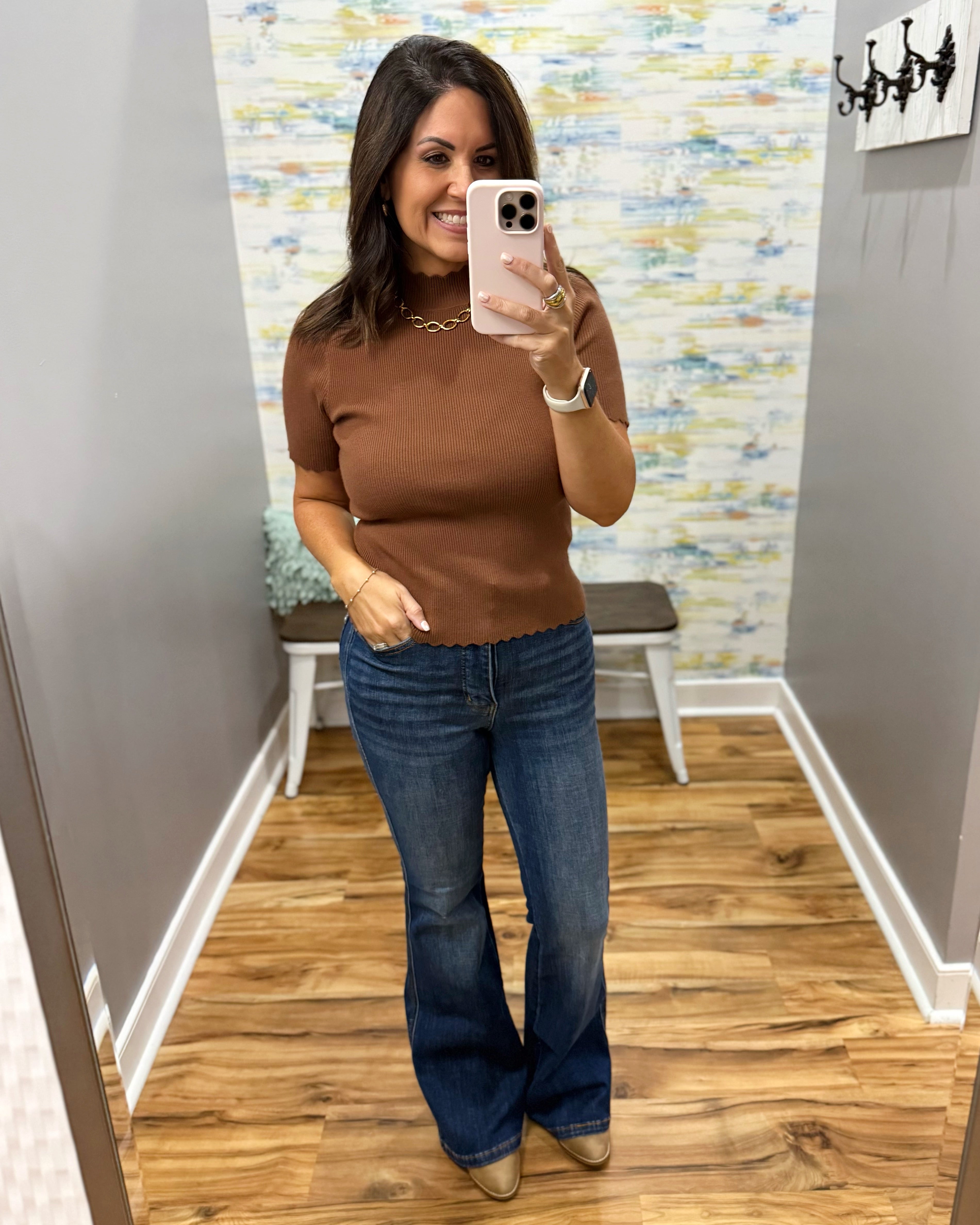 A woman takes a mirror selfie in a fitting room wearing a brown short-sleeve ribbed knit top and blue flare jeans.