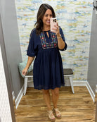 Woman taking a mirror selfie wearing a navy dress with floral embroidery in a room with patterned wallpaper.