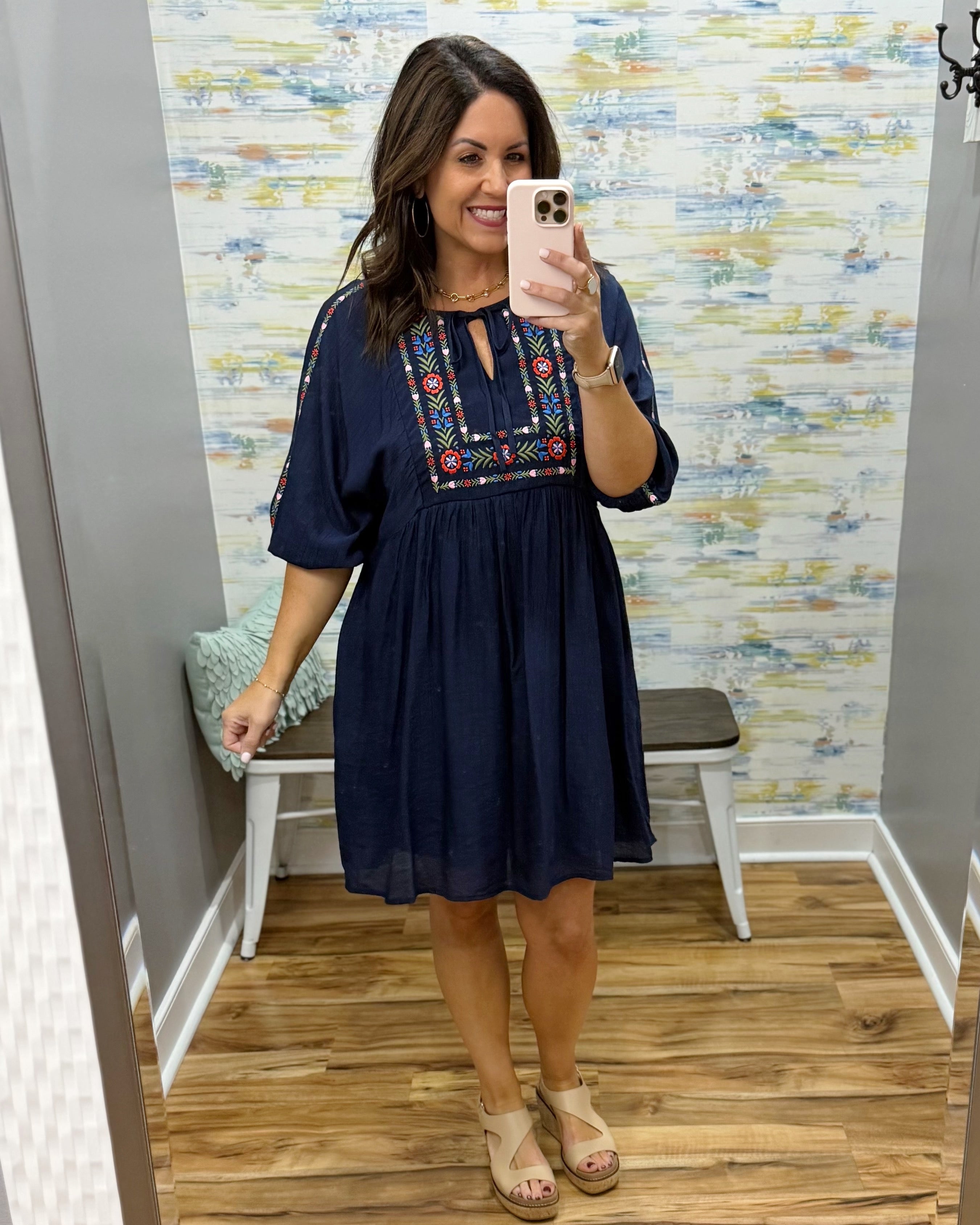 Woman taking a mirror selfie wearing a navy dress with floral embroidery in a room with patterned wallpaper.