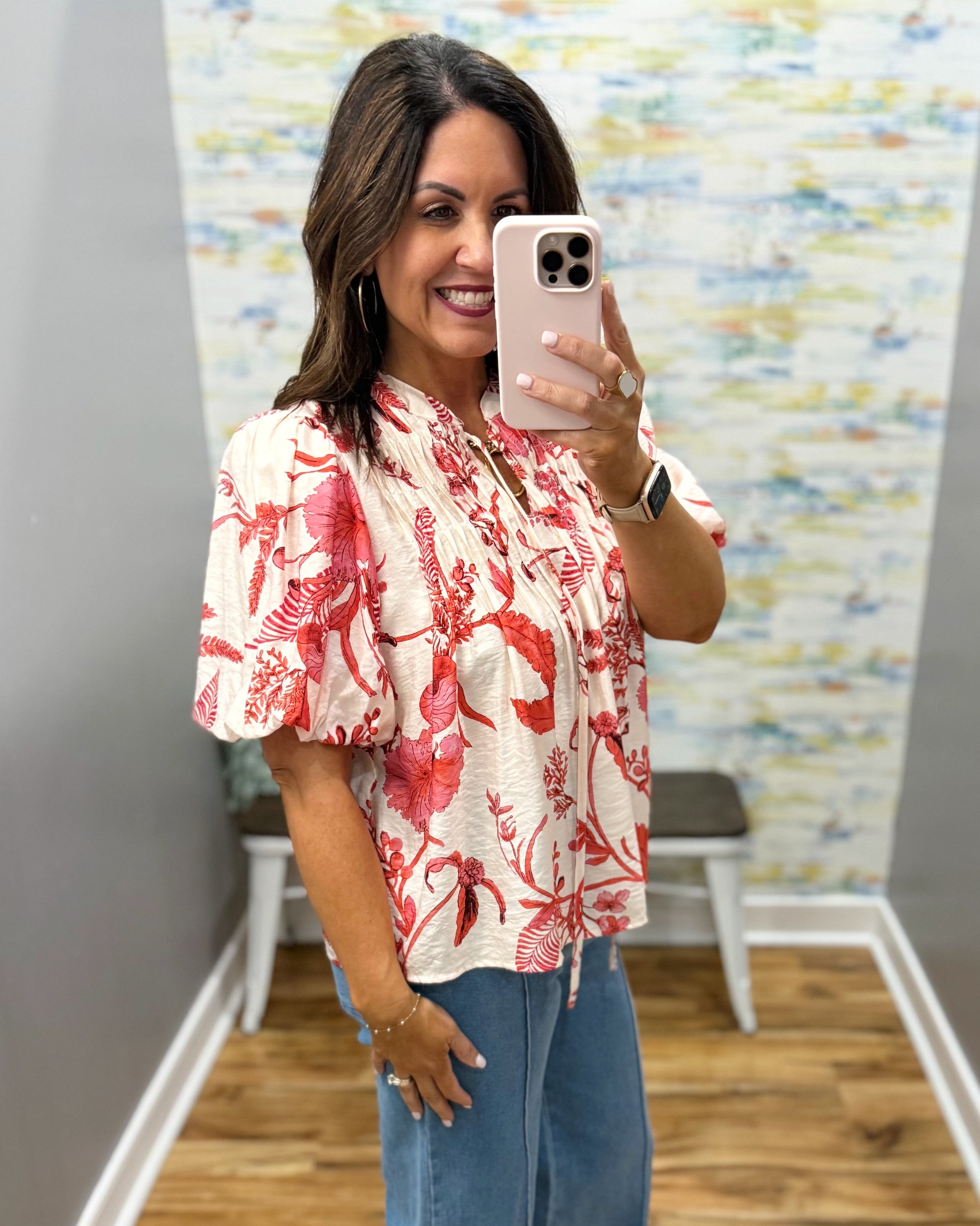 Woman taking a mirror selfie wearing a red floral blouse and blue jeans in a room with colorful wall art.