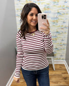 A woman taking a mirror selfie wearing a long-sleeve crewneck top with horizontal burgundy and white stripes, paired with dark blue jeans.