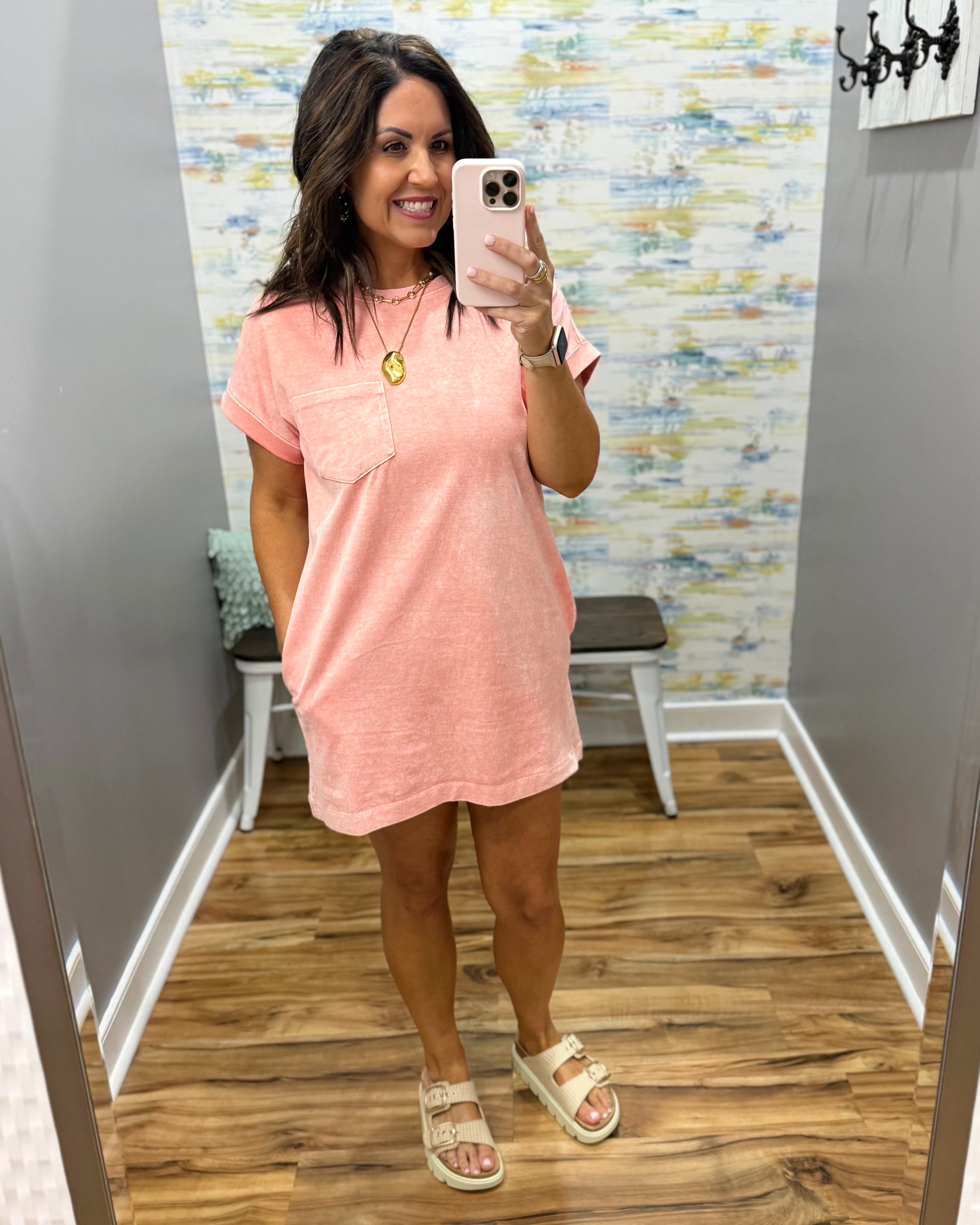 Woman taking a mirror selfie wearing a pink dress in a room with patterned wallpaper.