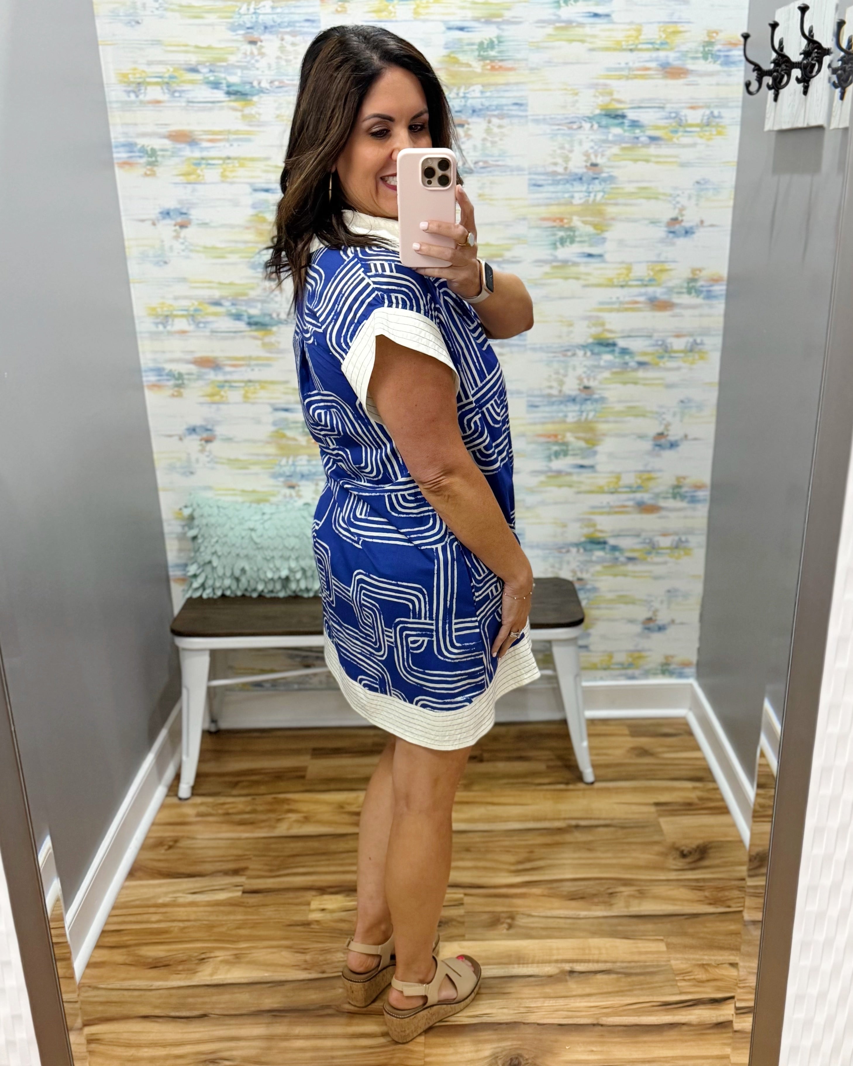 Woman taking a mirror selfie wearing a blue and white patterned dress in a room with a textured wall.