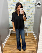 A woman takes a mirror selfie wearing a black short-sleeve pleated blouse and blue flare jeans in a fitting room with wood floors and a patterned wall.