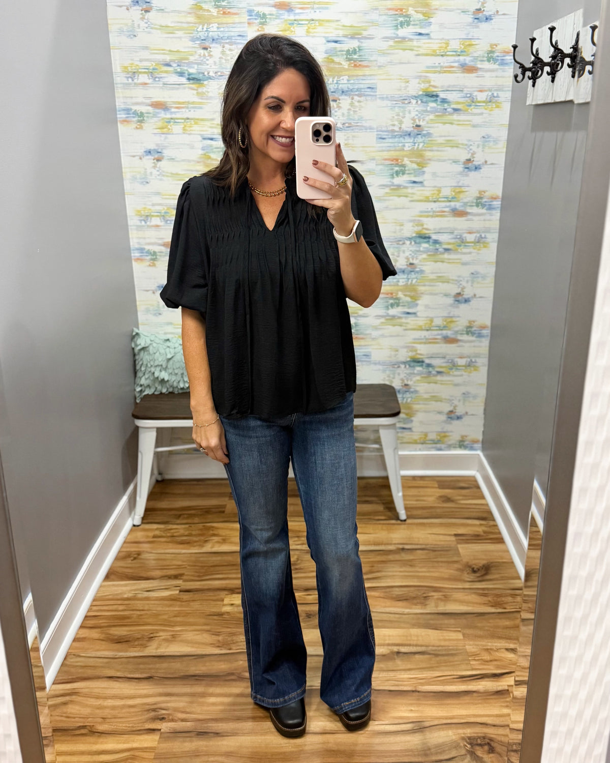 A woman takes a mirror selfie wearing a black short-sleeve pleated blouse and blue flare jeans in a fitting room with wood floors and a patterned wall.