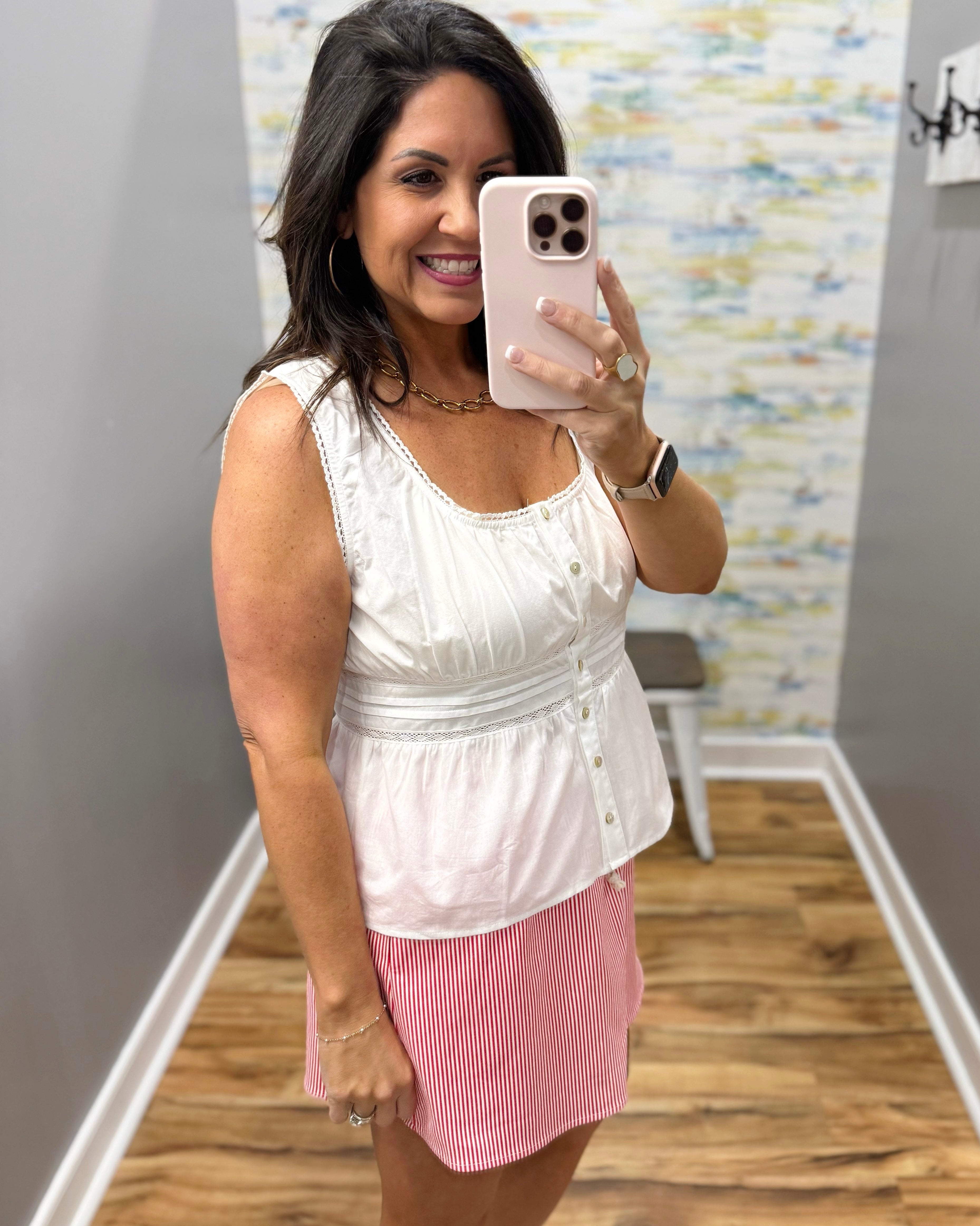 Woman taking a mirror selfie wearing a white sleeveless top and pink shorts.