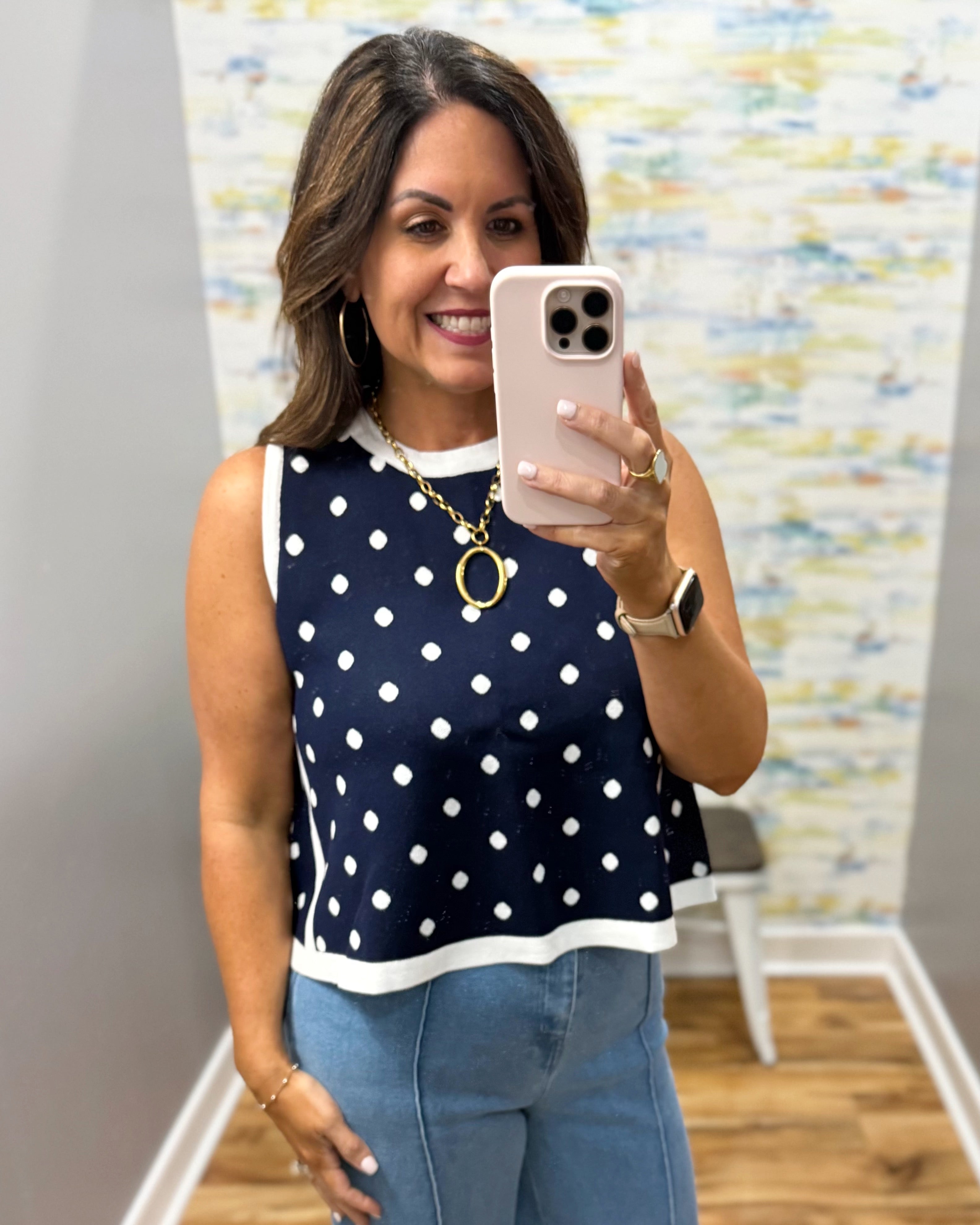 Woman taking a mirror selfie wearing a navy blue top with white polka dots and blue jeans.