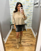 A woman takes a mirror selfie in a fitting room wearing a beige knit sweater, a black denim mini skirt, and beige slouchy knee-high boots.