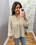 A woman takes a mirror selfie wearing a beige and black vertically striped long-sleeve blouse with a split neckline and gold pendant necklace, paired with blue jeans.
