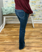A person wearing mica dark blue flare jeans and a burgundy top, standing on a wood floor in front of a white bench and a colorful wall.