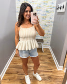 Woman taking a mirror selfie wearing a beige peplum top and denim shorts in a room with a colorful wall.
