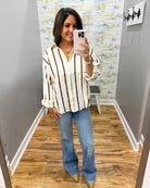 A woman takes a mirror selfie in a fitting room wearing a cream and brown vertically striped button-down shirt, blue flare jeans, and metallic gold pointed-toe flats.