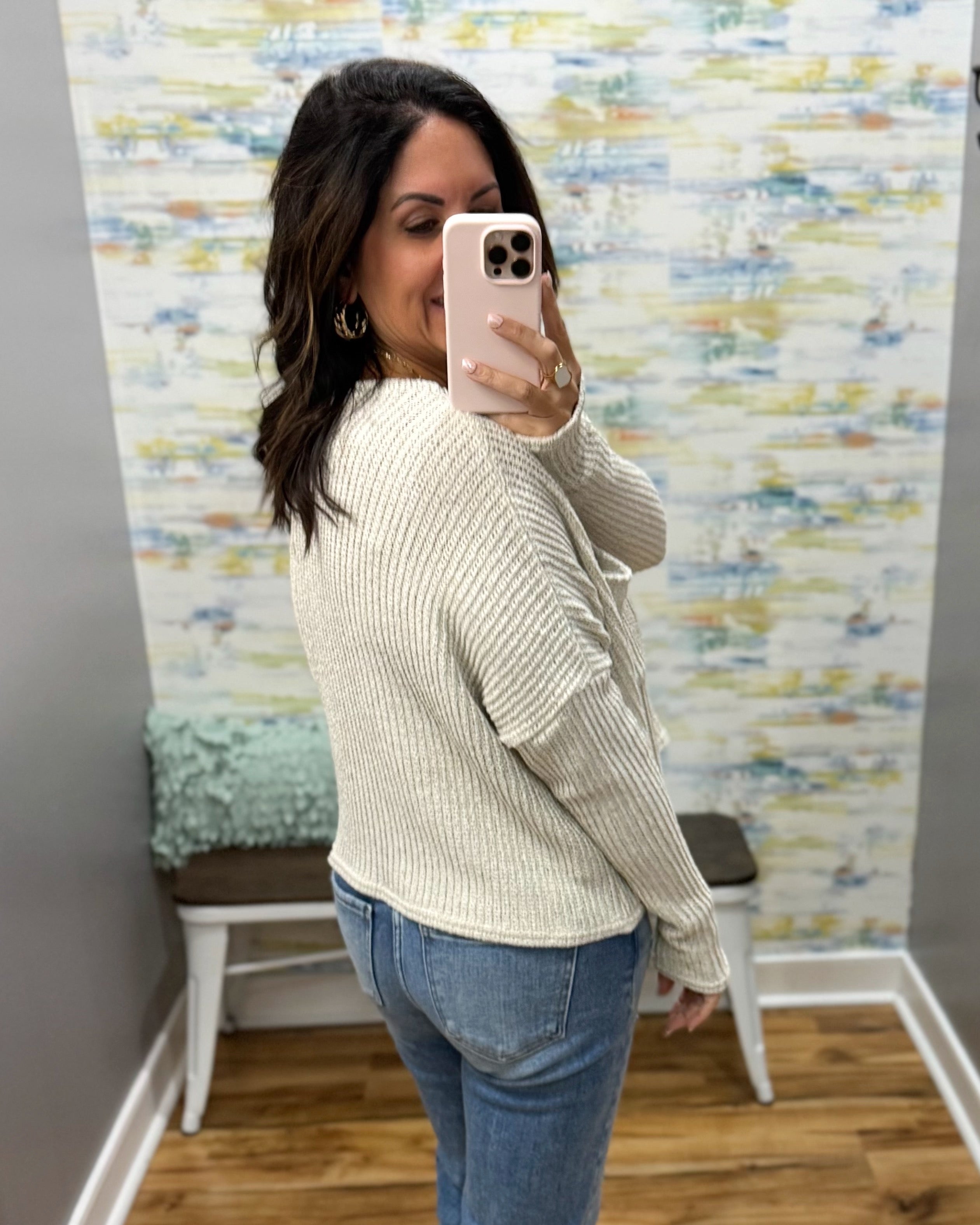 A woman takes a mirror selfie in a fitting room wearing a cream ribbed knit sweater and blue jeans, with a patterned wallpaper background.