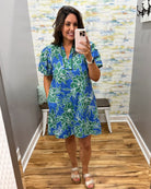 Woman taking a mirror selfie wearing a blue and green floral dress in a room with a patterned wall.