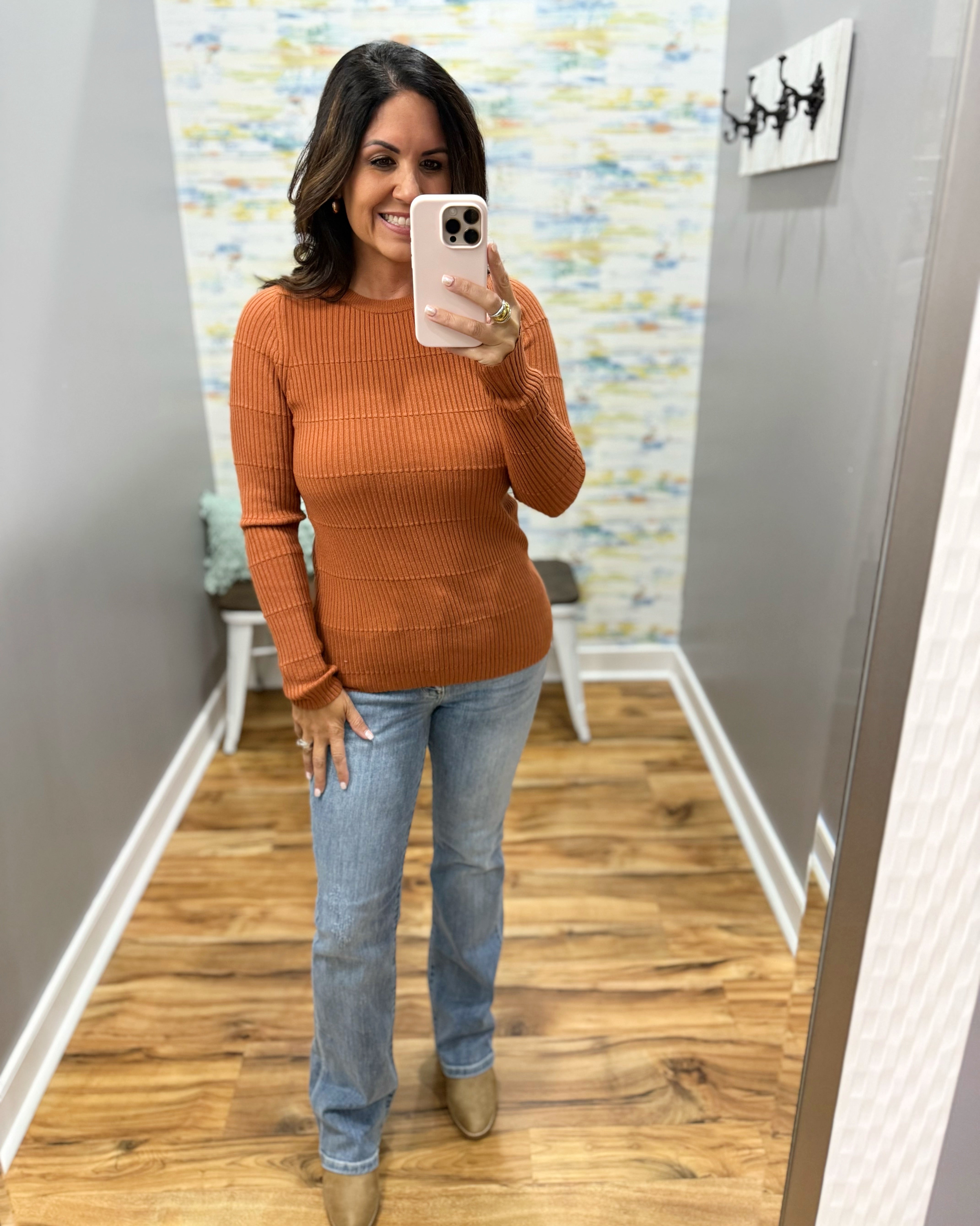 A woman takes a mirror selfie wearing a ribbed long-sleeve crewneck sweater in a burnt orange color, paired with light-wash blue jeans and tan boots, in a fitting room with wood floors.