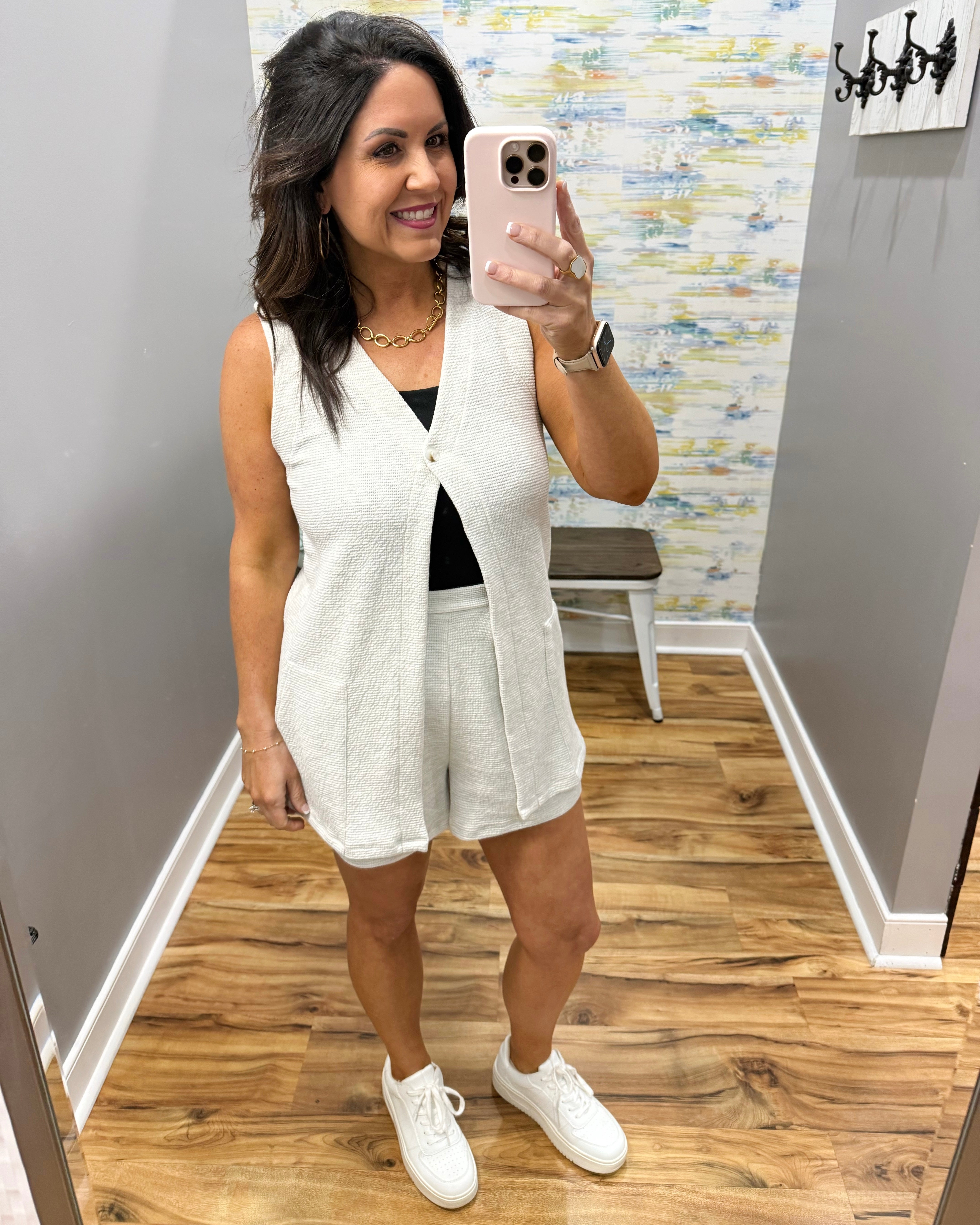 Woman taking a mirror selfie wearing a white sleeveless top and shorts with white sneakers.