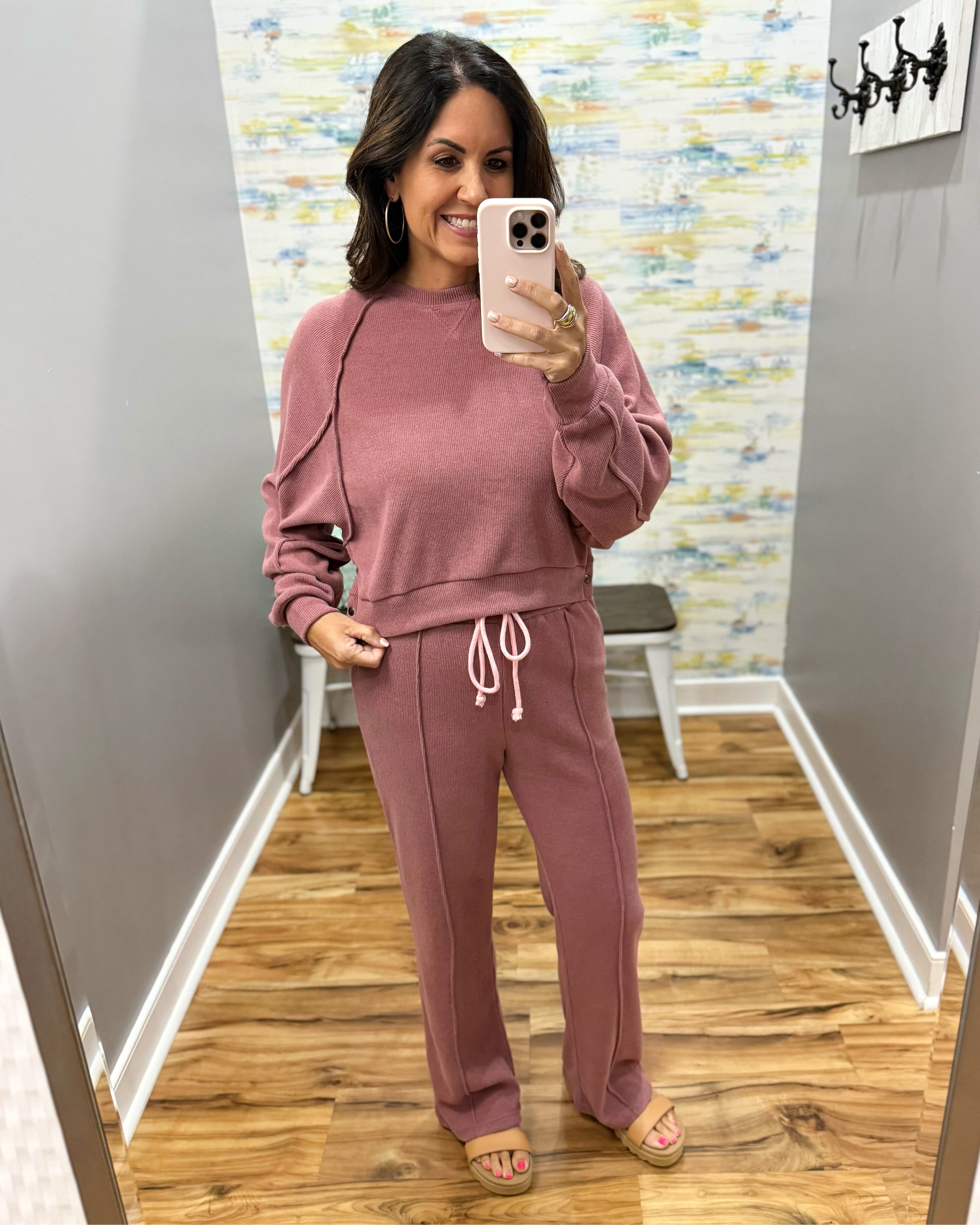 A woman takes a mirror selfie wearing a matching dusty rose sweatshirt and sweatpants lounge set with white drawstrings, standing in a fitting room with wood floors and gray walls.