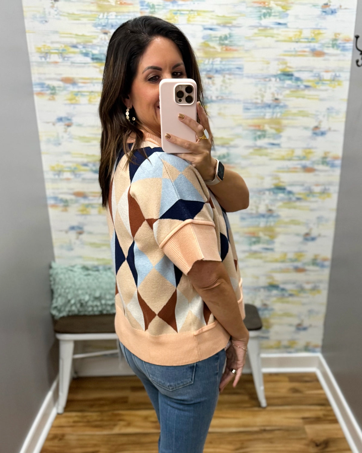 A woman takes a mirror selfie wearing a short-sleeve knit sweater with a geometric diamond pattern in peach, beige, light blue, and navy, paired with blue jeans.