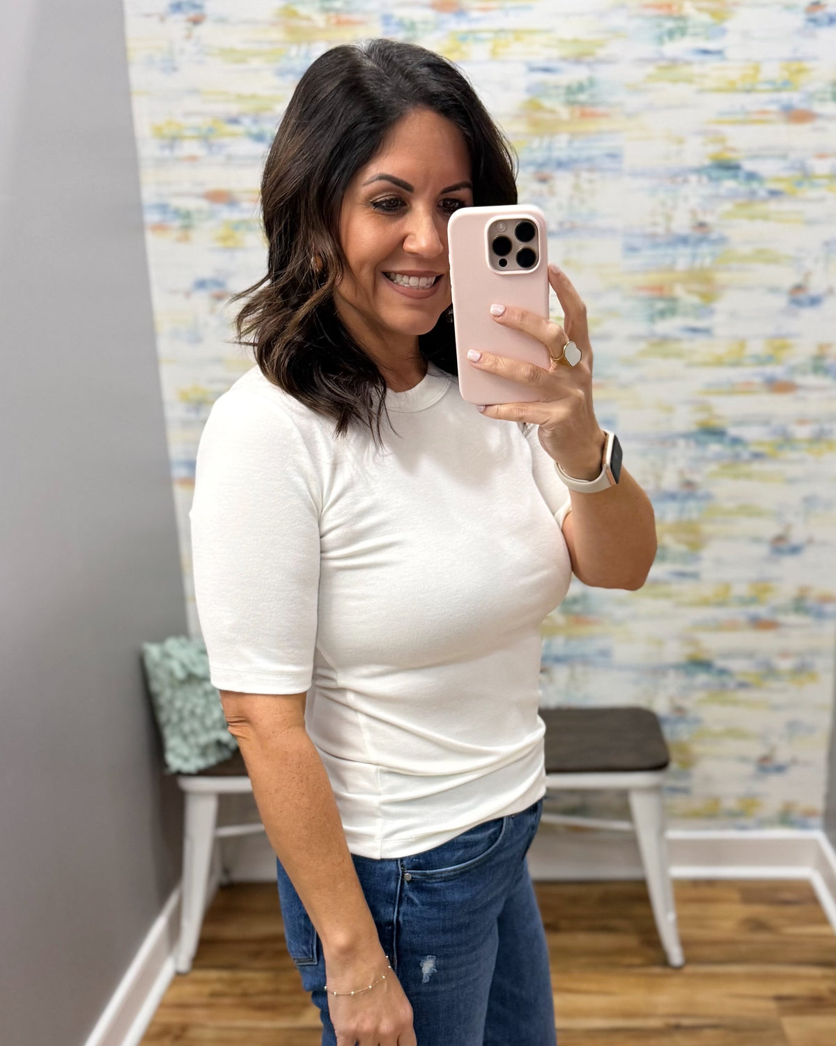 A woman taking a mirror selfie wearing a fitted white short-sleeve crewneck t-shirt and blue jeans in a room with a patterned wall.