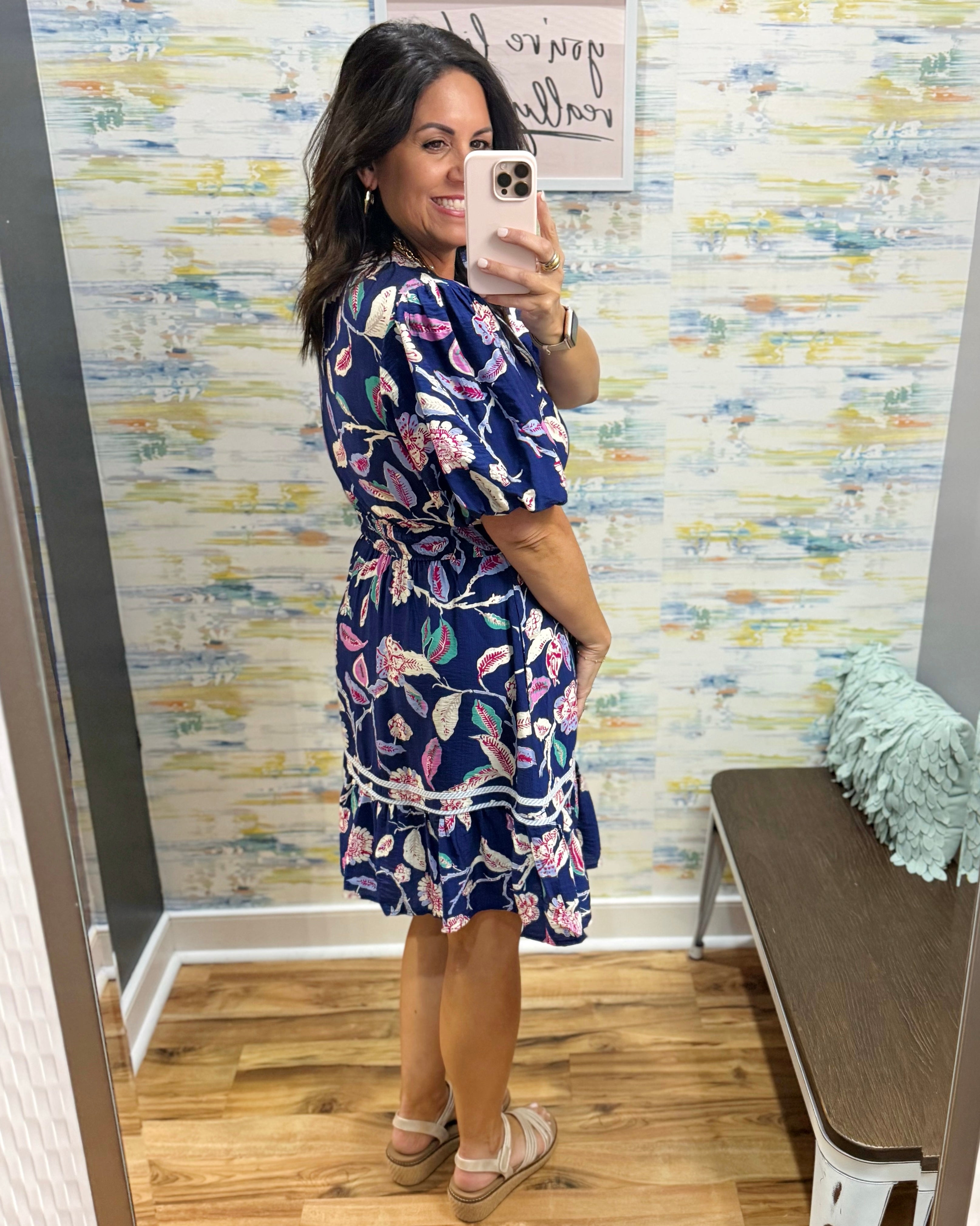 Woman taking a mirror selfie wearing a blue floral dress in a room with colorful wallpaper.