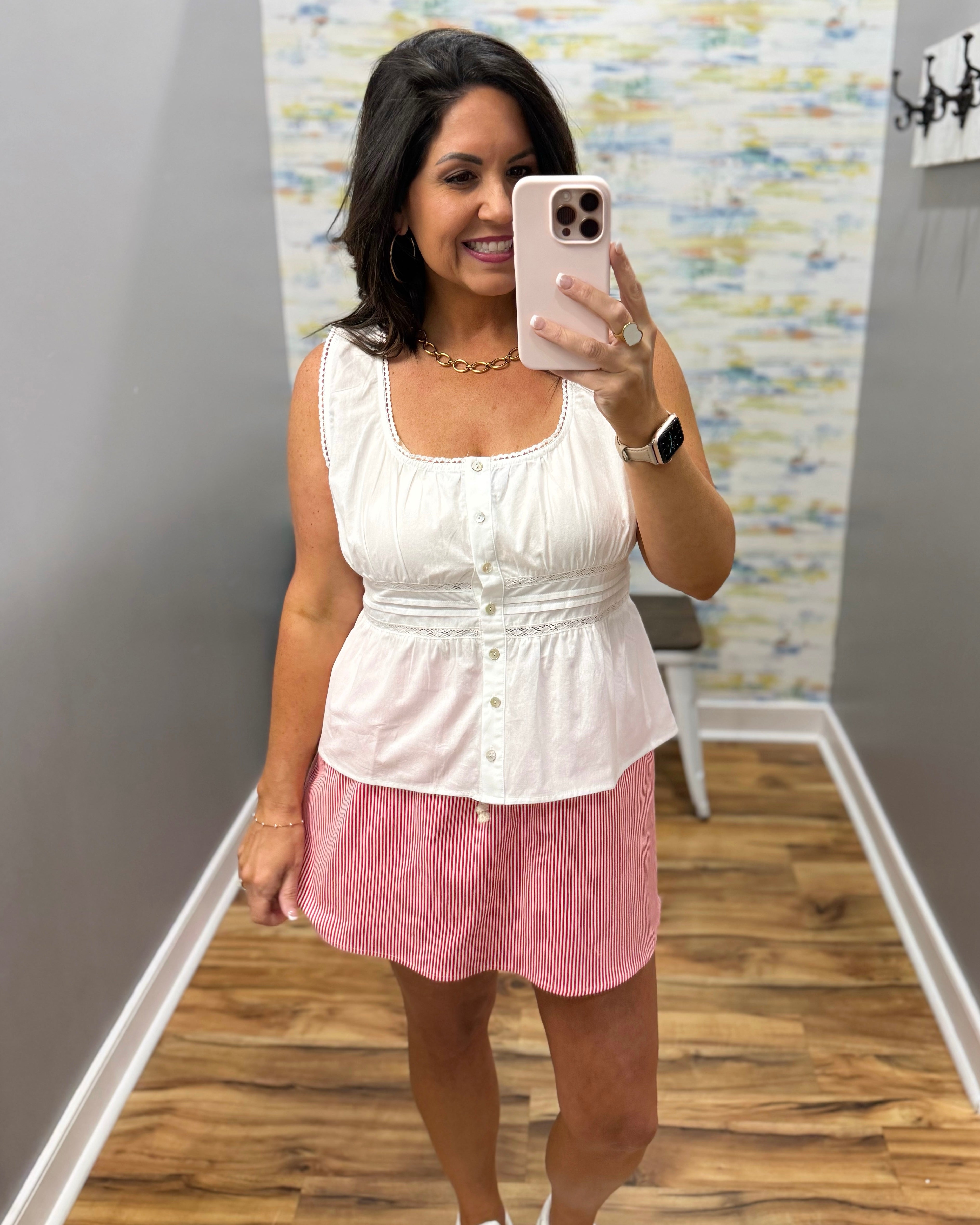Woman taking a mirror selfie wearing a white top and pink skirt in a room with a patterned wall.