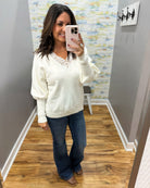 A woman takes a mirror selfie wearing a cream ribbed knit sweater with a lace V-neck detail, paired with dark blue flare jeans and brown shoes, in an indoor hallway setting.