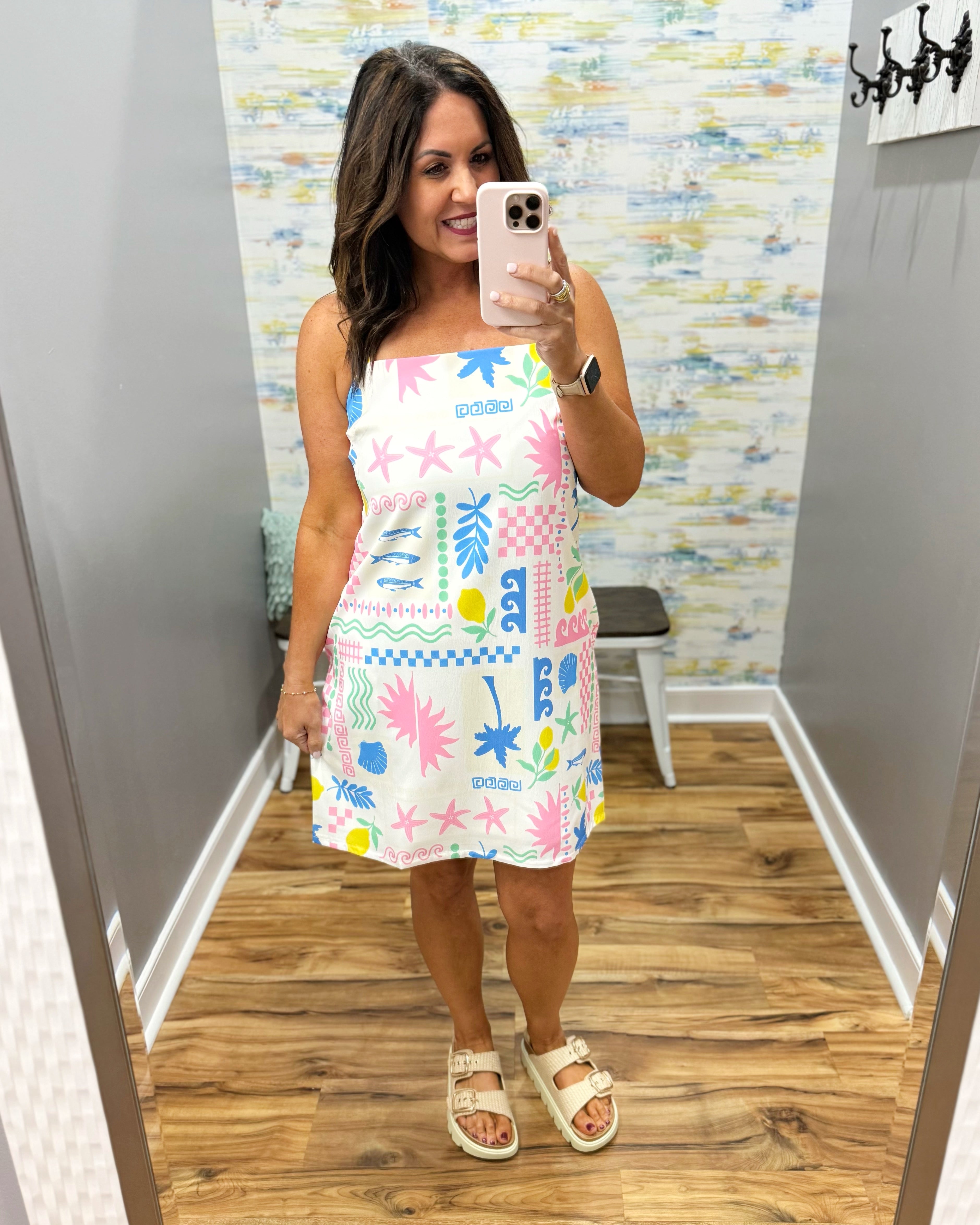 Woman taking a mirror selfie wearing a colorful dress with a pattern of shapes and letters.