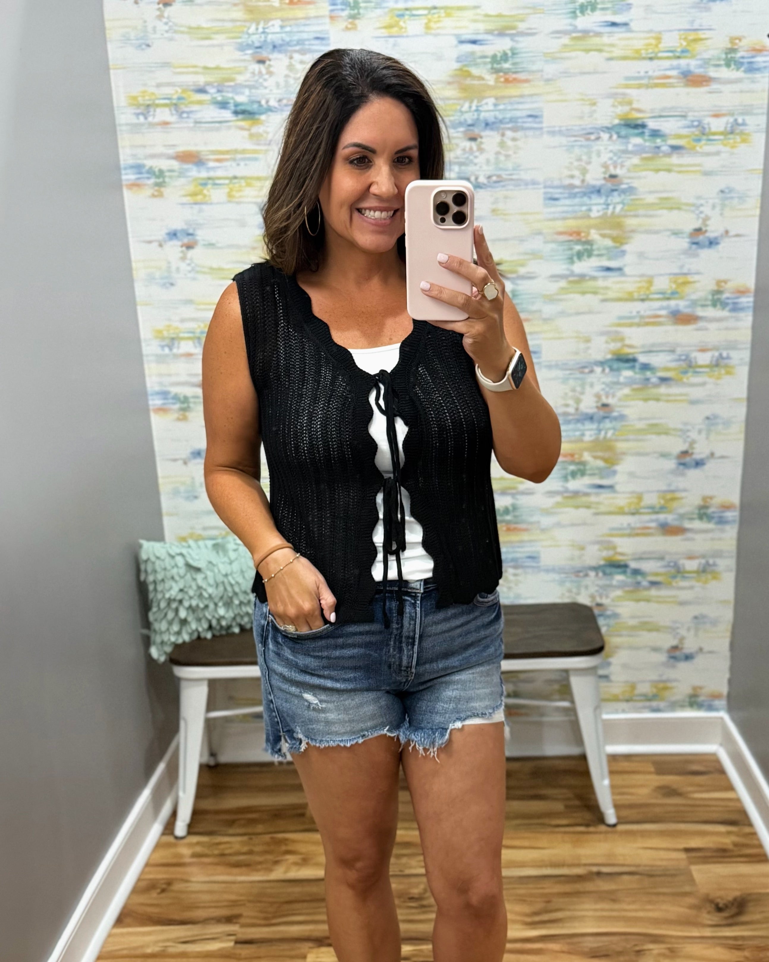 Woman taking a mirror selfie wearing a black sleeveless top and denim shorts in a room with colorful wall paper.