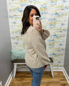 A woman takes a mirror selfie wearing a beige and brown vertically striped long-sleeve blouse with blue jeans, standing in front of a patterned wallpapered wall.