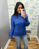 A woman takes a mirror selfie wearing a bright royal blue textured knit sweater and light-wash distressed jeans in a fitting room with a patterned wallpaper background.