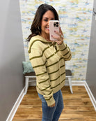 A woman takes a mirror selfie wearing a taupe and olive green striped knit hoodie sweater with blue jeans, standing in a room with wood floors and a colorful abstract wallpaper.
