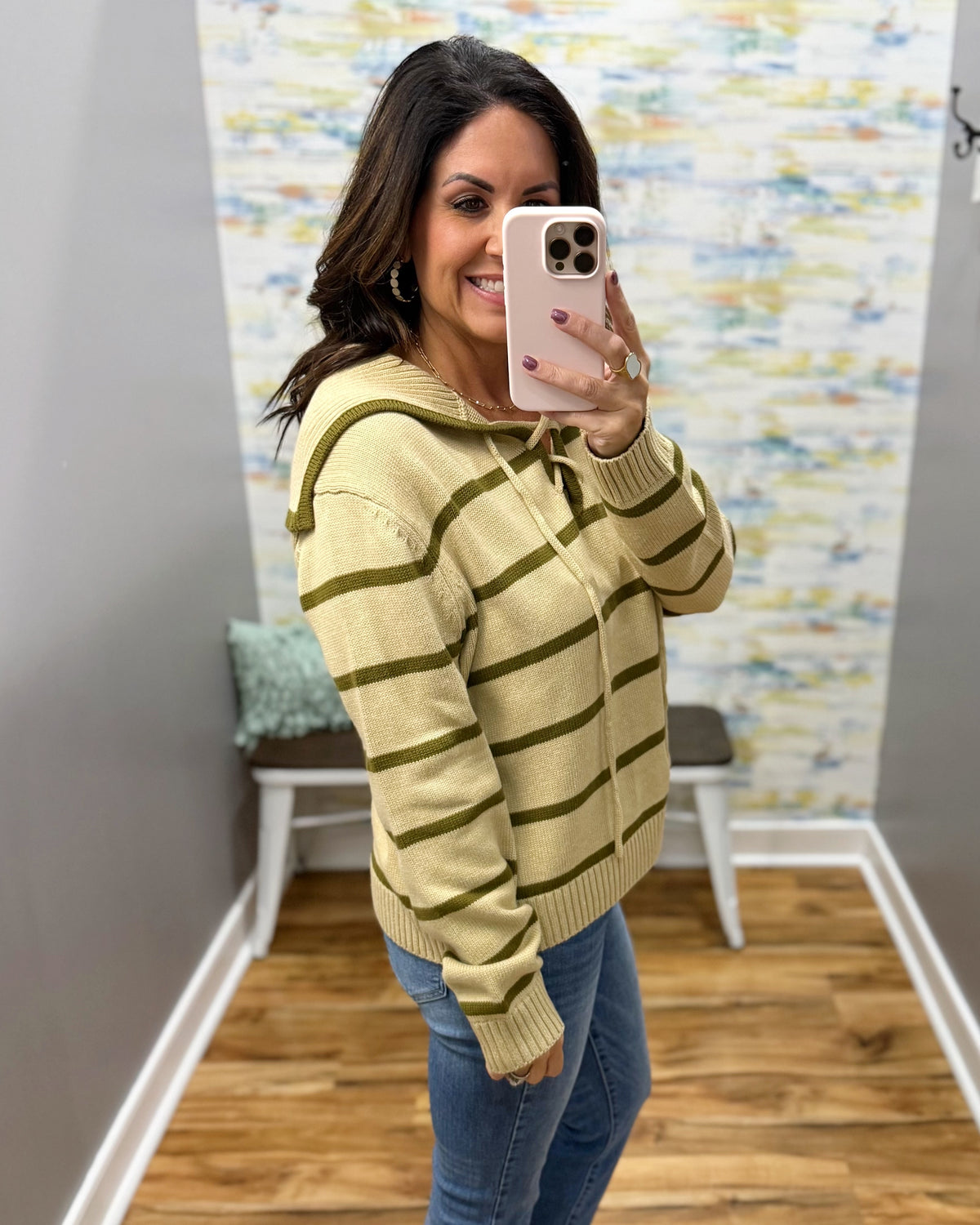 A woman takes a mirror selfie wearing a taupe and olive green striped knit hoodie sweater with blue jeans, standing in a room with wood floors and a colorful abstract wallpaper.