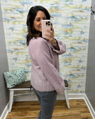 A woman takes a mirror selfie in a fitting room wearing a light pink knit sweater and gray pants, with a patterned wallpaper background and a bench with a pillow.