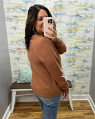 A woman takes a mirror selfie wearing a rust-brown knit sweater and blue jeans in front of a patterned wallpaper background.