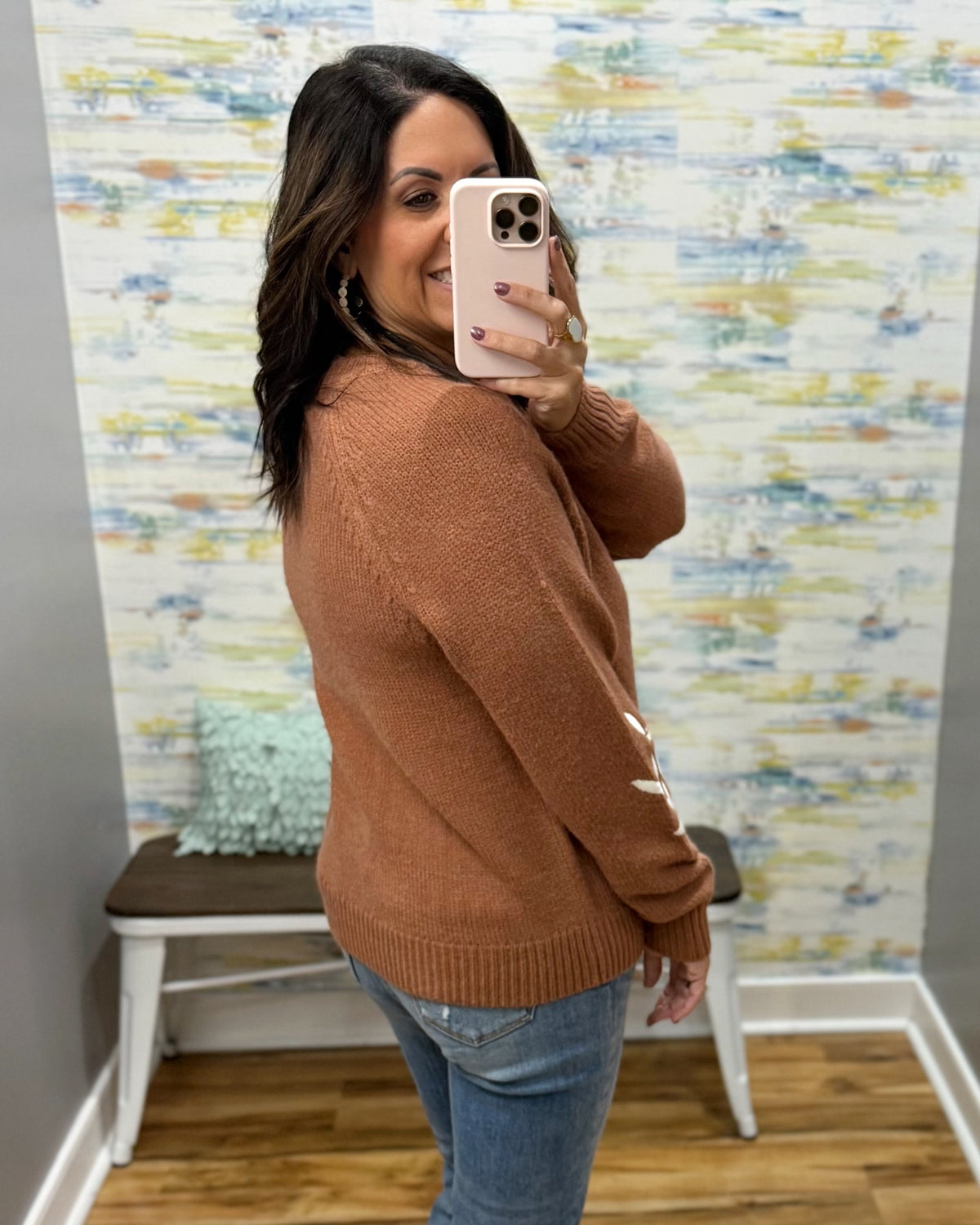 A woman takes a mirror selfie wearing a rust-brown knit sweater and blue jeans in front of a patterned wallpaper background.