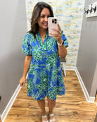 Woman taking a mirror selfie wearing a blue floral dress in a room with a patterned wall.