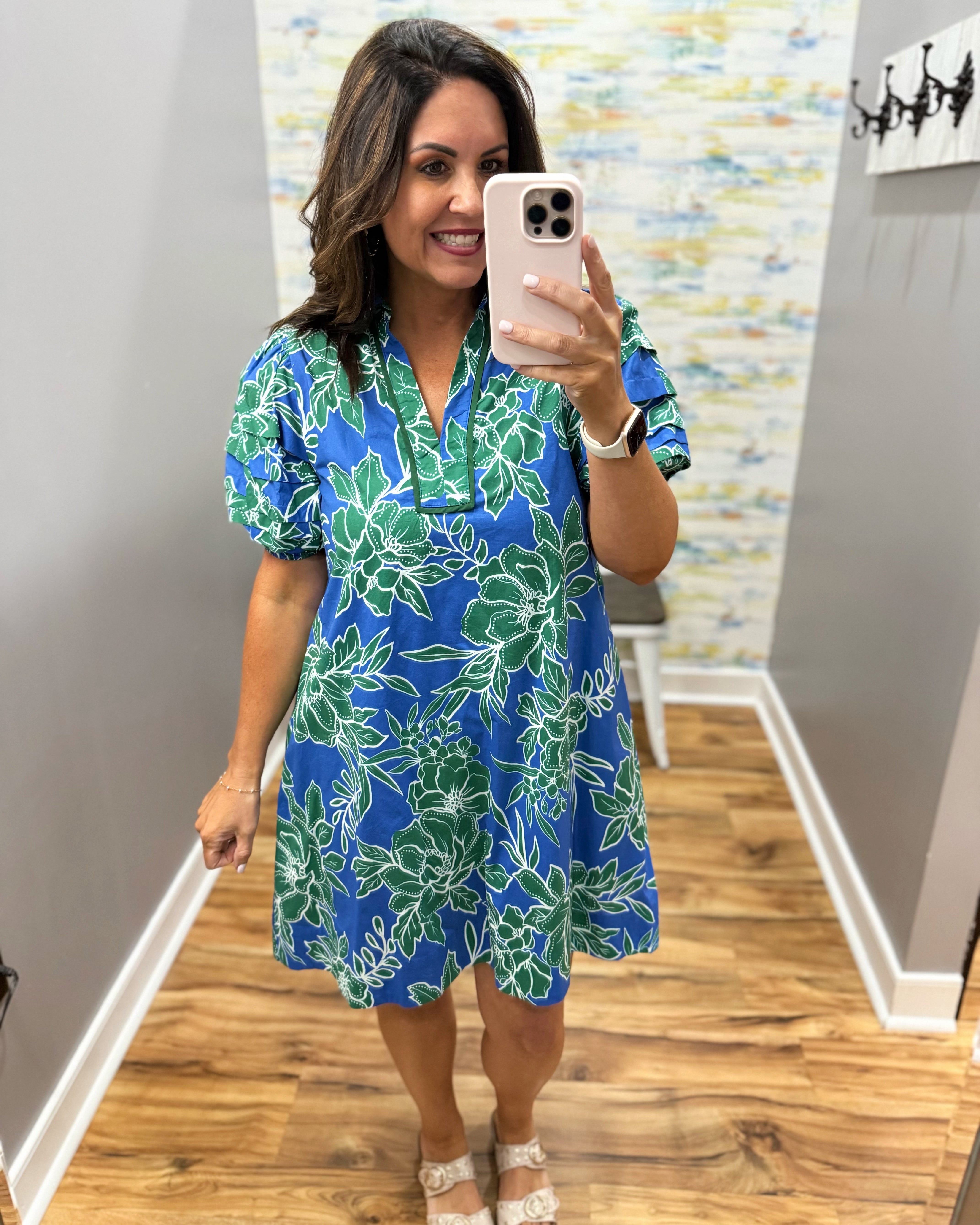 Woman taking a mirror selfie wearing a blue floral dress in a room with a patterned wall.